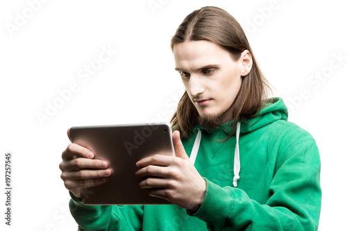 Portrait of young handsome beaded man in green hoodie looking at the digital tablet in his hands agains white background. Lifestyle, people and technology concept.