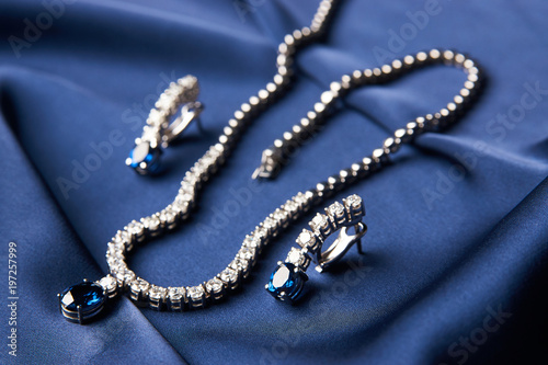 Women's platinum necklace and earrings with a diamond and blue precious sapphire stone on a silk blue background, close-up. Luxury female jewelry
