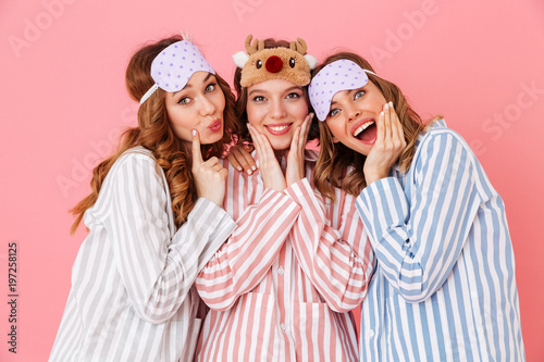Three beautiful young girls 20s wearing colorful striped pyjamas and sleeping masks having fun during girlish sleepover, isolated over pink background photo