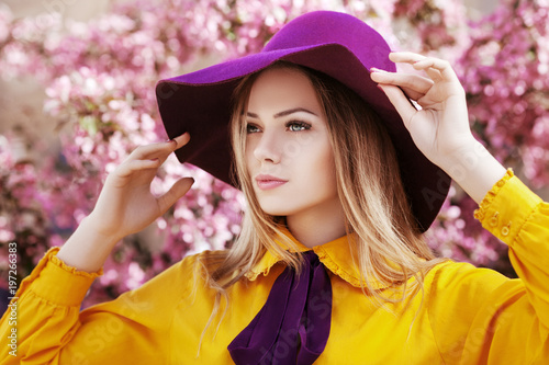 Outdoor close up portrait of young beautiful fashionable girl posing in street with blooming trees, wearing stylish wide-brimmed violet hat, yellow shirt, bow tie. Female spring fashion concept