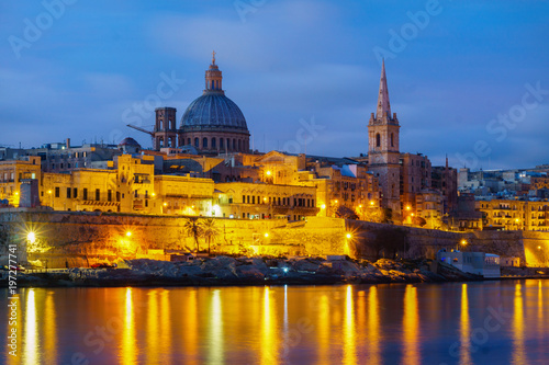 Night view of Valletta, capital of Malta. Beautiful spires and St. Pauls Cathedral and Charmelite Church skyline under blue night sky, lights reflected in the sea bay.