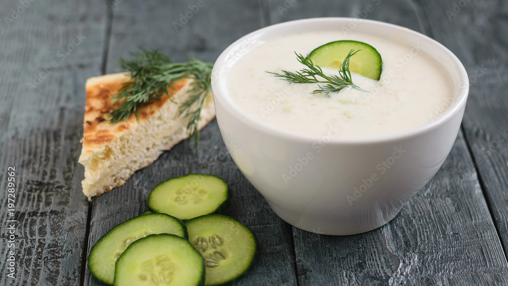 Freshly made tzatziki in a white bowl with cucumber and bread on a dark wooden table. Freshly made tzatziki in a white bowl with cucumber and bread on a dark wooden table.
