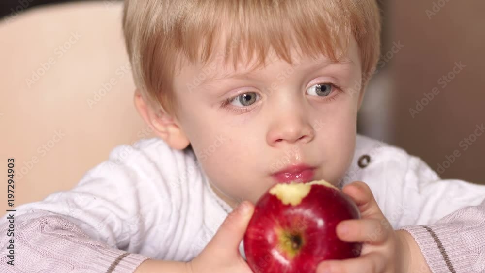 Close-up of a small blond boy eating a red apple sitting at a table at home, high resolution. Cute little child is biting red apple while sitting at table. Close-up of a small blond boy eating a red apple sitting at a table at home, high resolution. Cute little child is biting red apple while sitting at table.