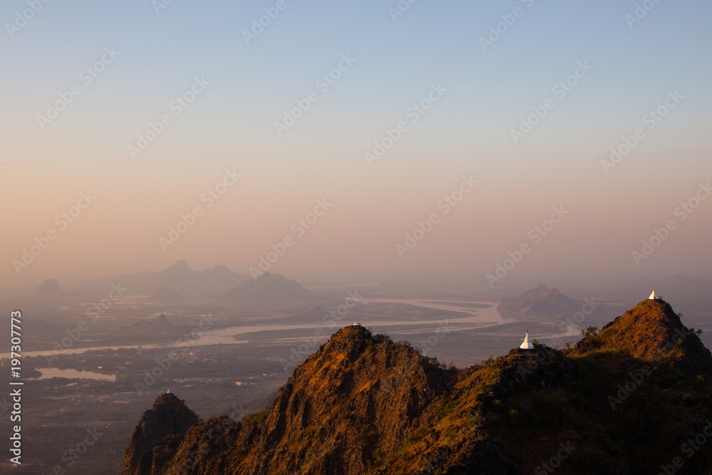 Scenic sunset view of white ancient buddhist stupas on peaks of textured mountains in yellow sun light. Misty valley with winding river and shadowy mountains background. Kayin State, Myanmar.