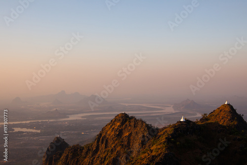 Scenic sunset view of white ancient buddhist stupas on peaks of textured mountains in yellow sun light. Misty valley with winding river and shadowy mountains background. Kayin State, Myanmar.