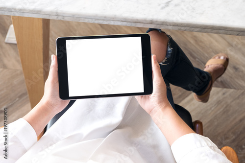 Top view mockup image of a woman sitting cross legged and holding black tablet pc with blank white desktop screen in cafe