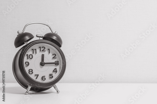 Closeup alarm clock for decorate show a quarter past twelve o'clock on white wood desk and cream wallpaper textured background in black and white tone with copy space