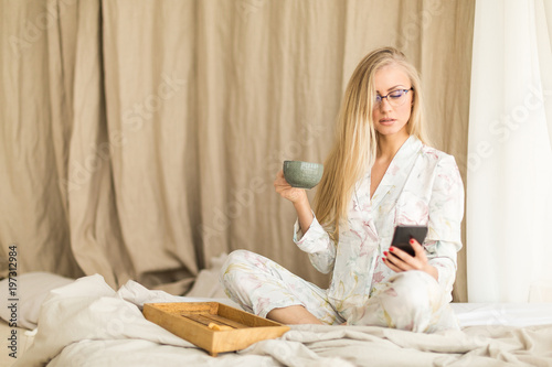 Young woman in glasses holding a cup of coffee and a mobile phone while in bed.