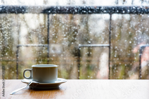 Tablou pe pânză A white espresso cup with a spoon on a wooden table in front of a glass window covered with rain drops
