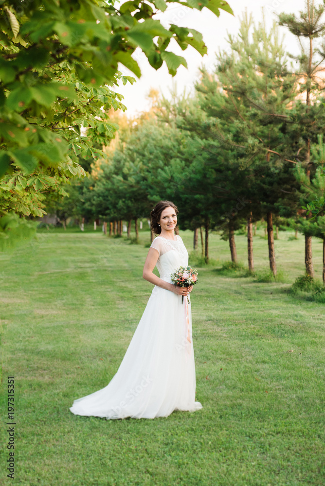 Chaming young girl in a wedding dress posing for a photo in a green garden. Beautiful bride in wedding dress outdoor in a field at sunset