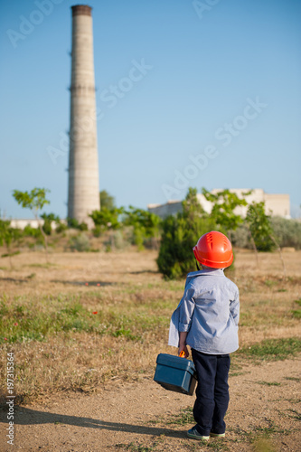 little preschooler kid in helmet with box of working tools in his hand looks at exhaust pipe of old plant