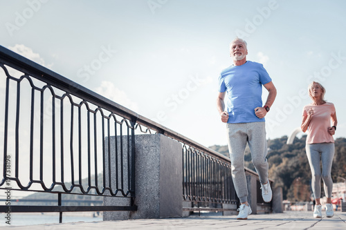 Fototapeta Naklejka Na Ścianę i Meble -  Follow me. Confident unshaken athletic man spending time on fresh air looking straight and running across the quay with a woman.