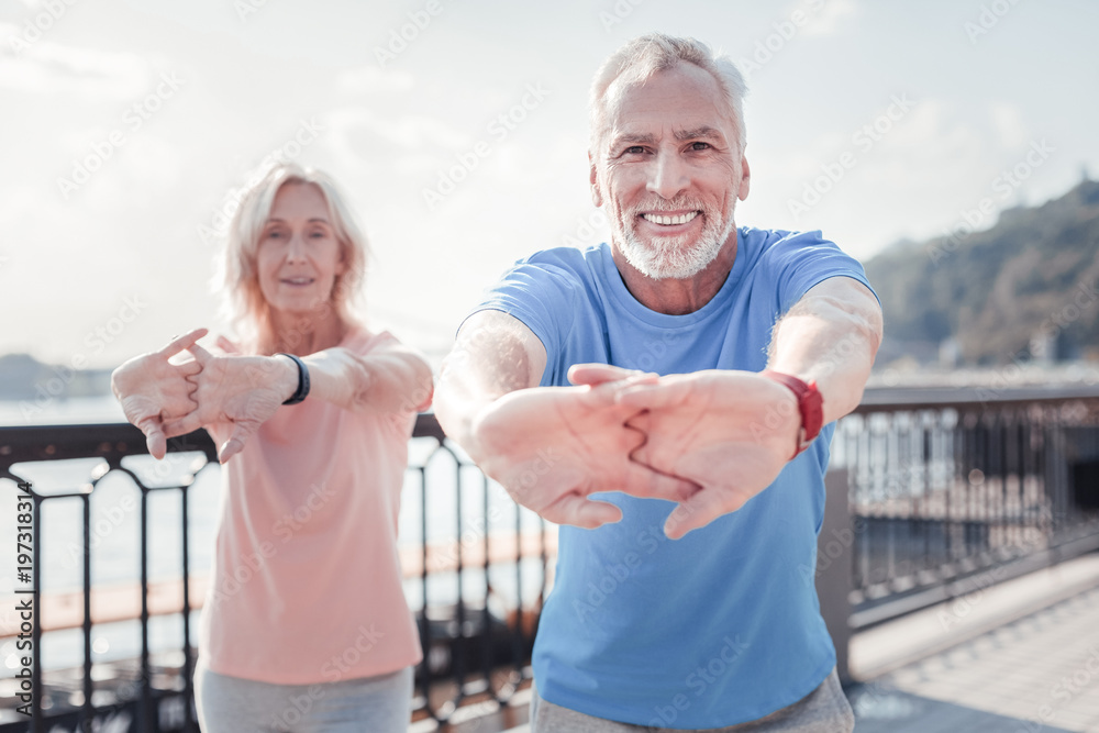 Together is better. Senior pleasant unshaken man standing with a woman smiling and training on the quay. Together is better. Senior pleasant unshaken man standing with a woman smiling and training on the quay.