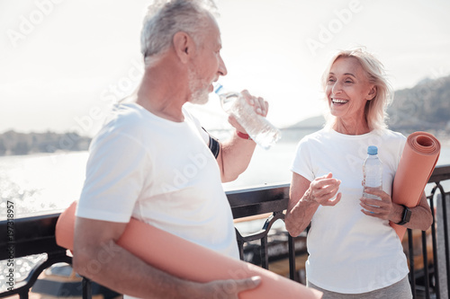 Good tome together. Happy senior sporting woman standing opposite man preparing for the training and drinking water.