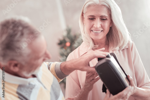 Lets look. Pleasant grey haired satisfied woman standing near her husband smiling and holding VR glasses.