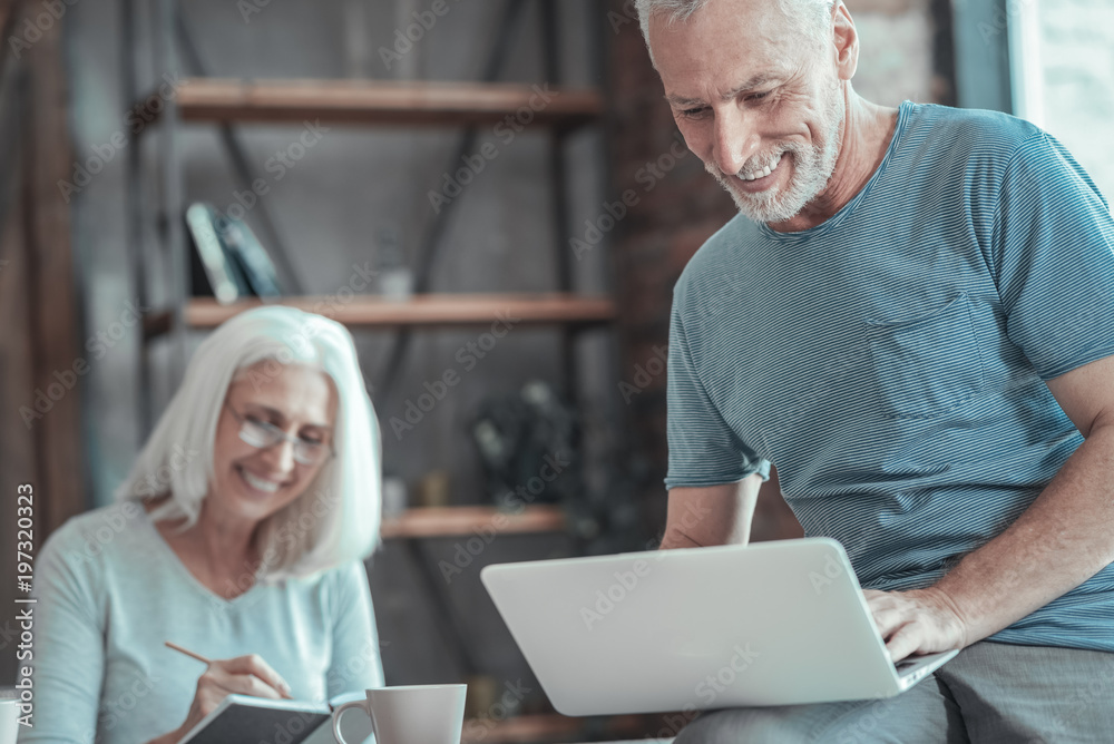 Interesting information. Satisfied unshaken aged man sitting on the table near a woman smiling and working with the laptop.