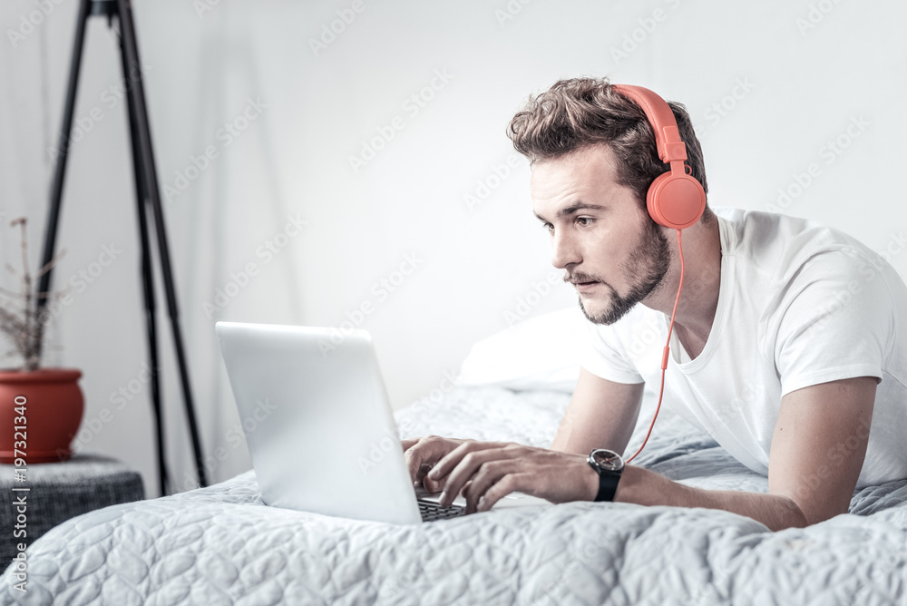Working at home. Nice pleasant handsome man lying on the bed and typing on the laptop while listening to music Working at home. Nice pleasant handsome man lying on the bed and typing on the laptop while listening to music