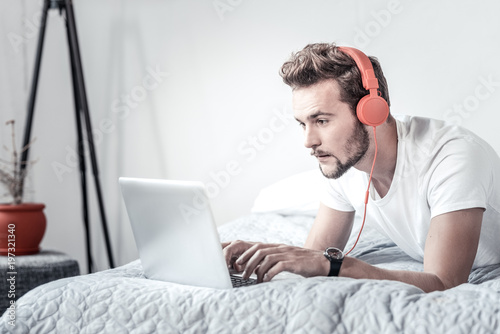 Working at home. Nice pleasant handsome man lying on the bed and typing on the laptop while listening to music © Viacheslav Yakobchuk