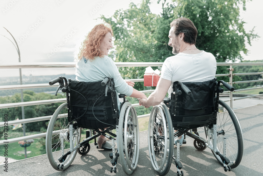 Just take it. Charming female person holding hand of her man and expressing positivity while being on her wheelchair