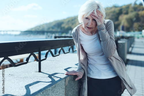Need to take my pills. Exhausted older lady leaning on a barrier and touching her forehead while suffering from a terrible headache outdoors. photo