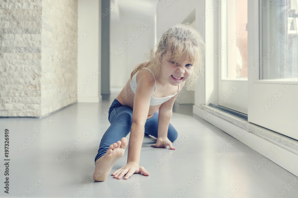Little blond girl in white top and blue leggings stretches a twine in the room near the window Little blond girl in white top and blue leggings stretches a twine in the room near the window