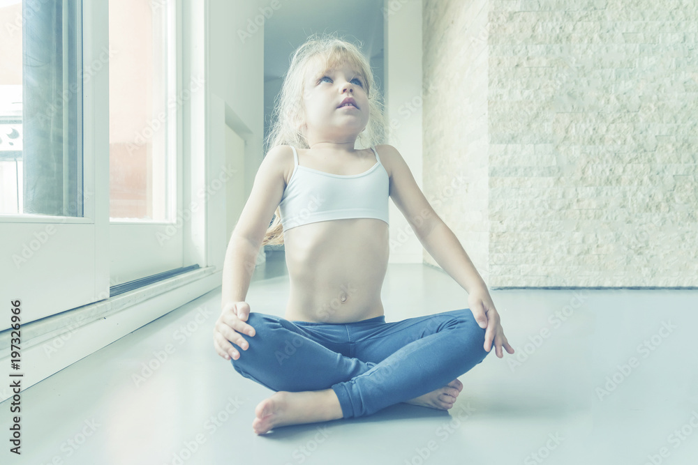 Little blond girl in white top and blue leggings doing a yoga exercise in the room near the window Little blond girl in white top and blue leggings doing a yoga exercise in the room near the window