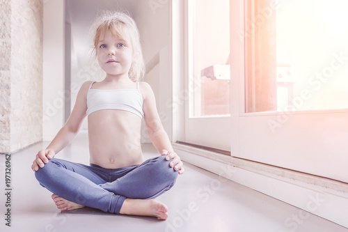 Little blond girl in white top and blue leggings doing a yoga exercise in the sunny room