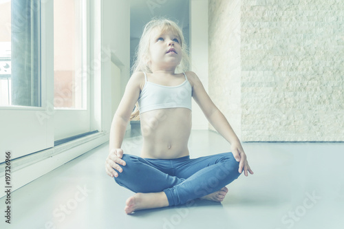 Little blond girl in white top and blue leggings doing a yoga exercise in the room near the window