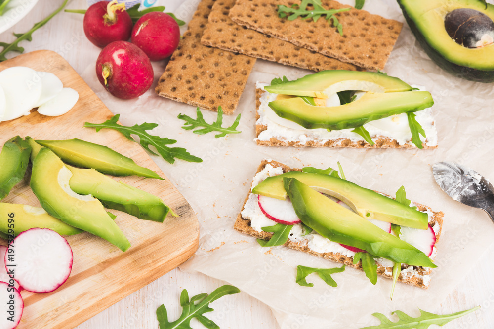 Process of making sandwiches whith green avocado. Red garden radish, arugula, cream cheese, eggs and rye bread crusts. Healthy food on wooden chopping board. 