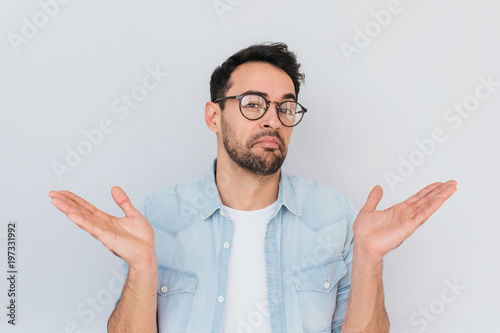 Portrait of uncertain young stylish stubble man with trendy round glasses wears demin blue shirt, shrugs shoulders being puzzled or confused. Caucasian unsure male make gestures doubtfully with hands. photo