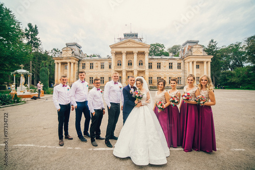 Newlywed couple, bridesmaids & groomsmen having fun outdoors. Bride and groom with friends near old medieval castle. Wedding moment of newlyweds.