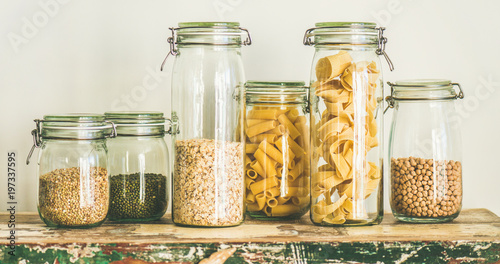 Various uncooked cereals, grains, beans and pasta for healthy cooking in glass jars on rustic table, white background, wide composition. Clean eating, vegetarian, vegan, balanced dieting food concept