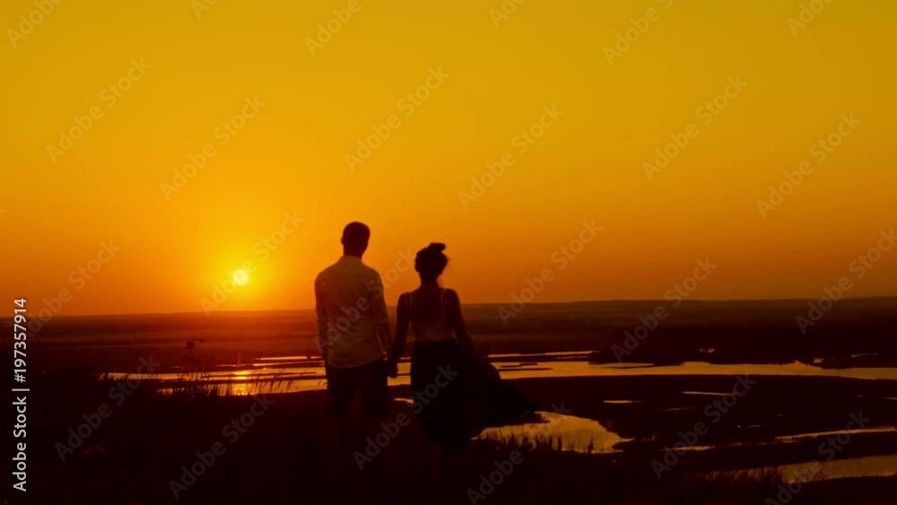 Loving couple at sunset - young man and beautiful girl stands at summer meadow and looking to sun, silhouette Loving couple at sunset - young man and beautiful girl stands at summer meadow and looking to sun, silhouette