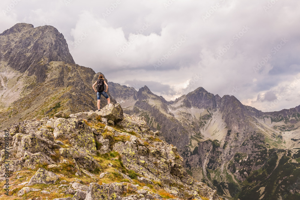 Fototapeta premium Surowy krajobraz górski, z ciemnymi chmurami. Kobieta obserwująca górską panoramę po wejściu na szczyt. Tatry Wysokie w Słowacji.