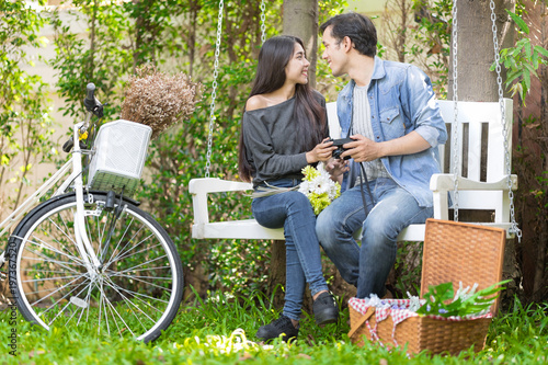 Sweet Love Couple are sitting on swings in the garden and holding flowers on Weekend Having Lunch and Chill out with Beautiful Nature.