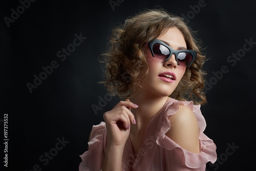 Young attractive model is looking at side,posing in front of camera and wearing fashionable sunglasses with nice pink dress. Woman is having accurate manicure and day make up. Dark grey background. photo