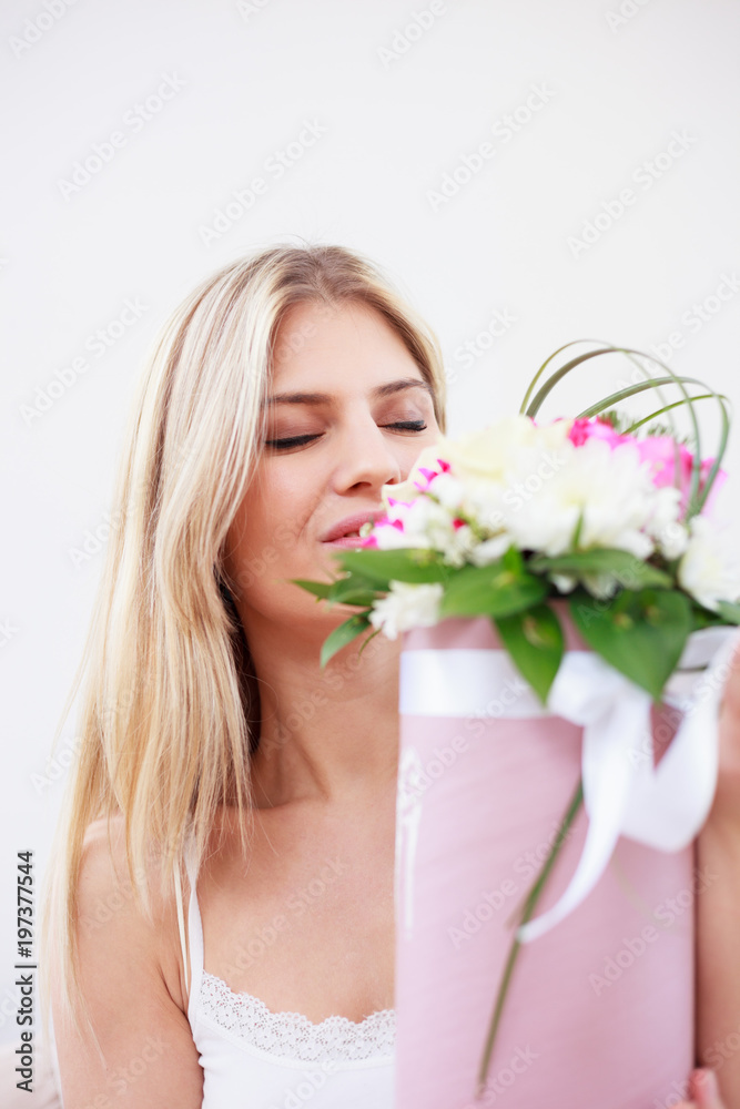 Beautiful happy young woman smelling a bouquet of violet and white flowers she just received. Relaxing with closed eyes on a sofa in pajamas in a brightly lit room.