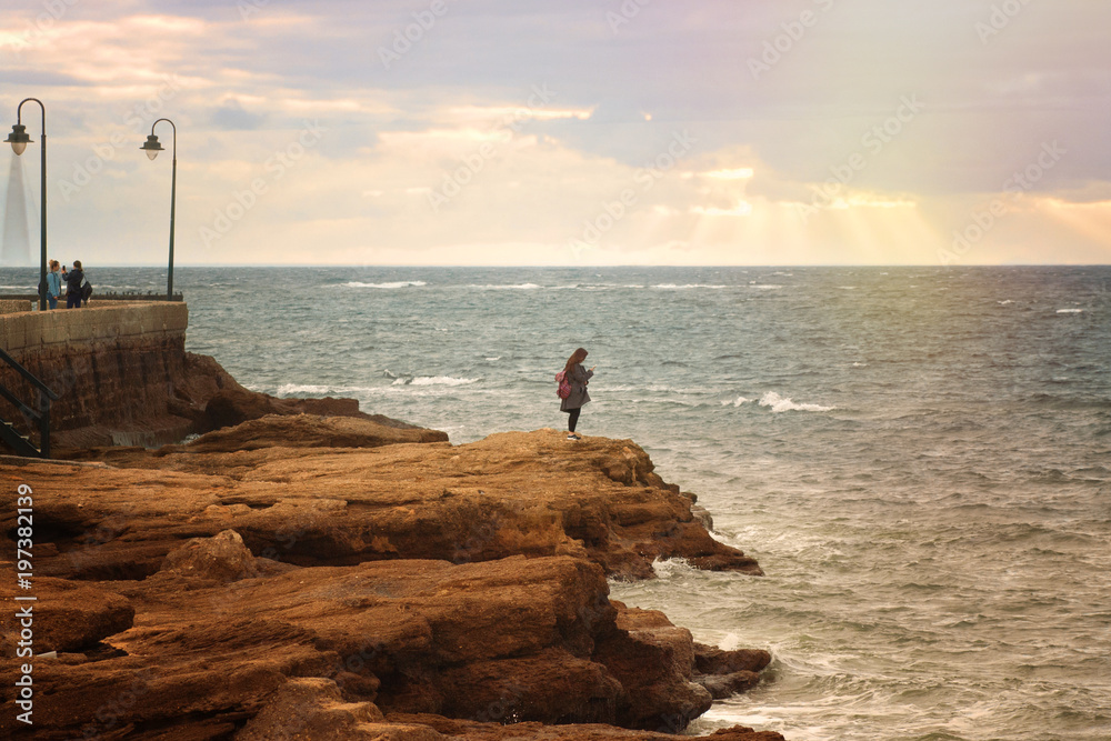 the girl stands on the huge rocks and looks at the sea holding the phone