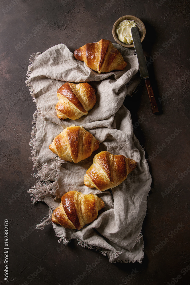 Fresh baked traditional croissant on linen cloth with butter and vintage knife over dark brown texture background. Top view, space