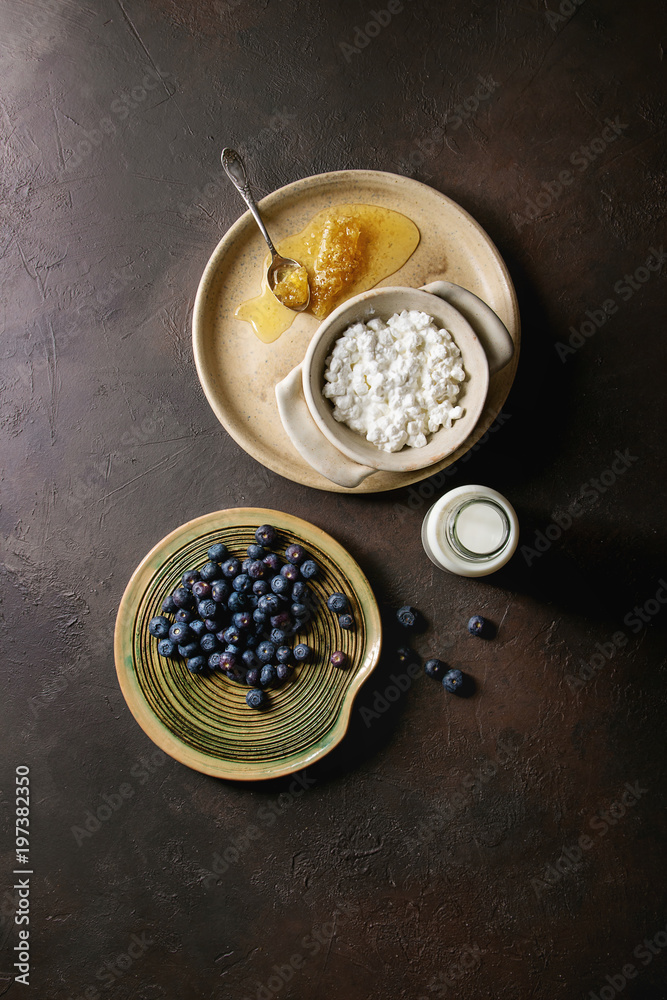 Ceramic bowl of homemade cottage cheese served with blueberries, bottle of milk and honeycombs over dark brown texture background. Top view, space
