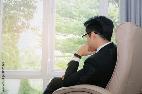 side view of young handsome business man with eyeglasses sitting on sofa chair in modern office with green garden nature background from window, savings, investment, finance and banking concept photo