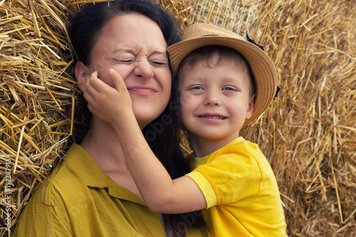 Happy family. A young woman with dark hair hugs her son in a hat against a background of hay. Mom and son. © AnastazjaSoroka