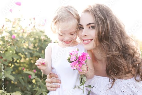 Beautiful blonde woman holding kid girl 3-4 year old holding flowers in meadow. Looking at camera. Motherhood. Summer season.