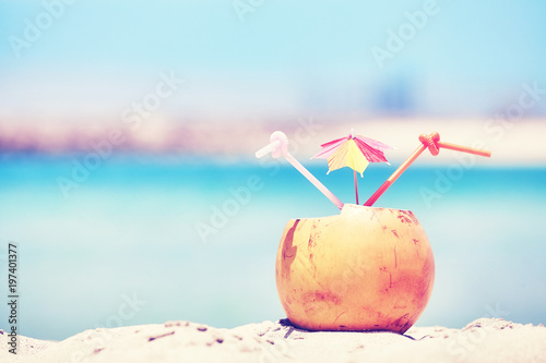 Retro toned picture of a coconut with two straws and colorful umbrella on a beach, summer holiday concept, selective focus.