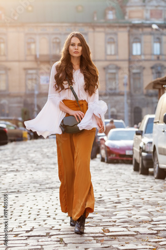 Outdoor full body fashion portrait of young beautiful woman wearing stylish yellow high-waisted wide-leg pants, white blouse, holding green leather bag. Model walking in street of european city