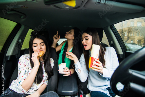 Three young women drive a car, speaking each other and eating a fast food in a car