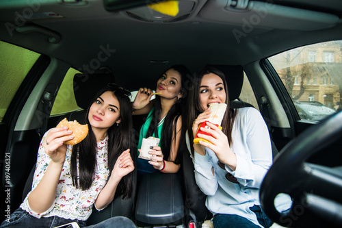 Three young women drive a car, speaking each other and eating a fast food in a car