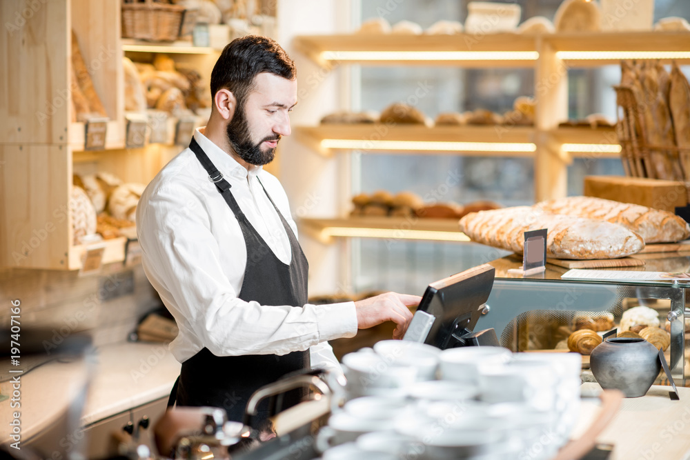 Handsome bread seller working with cash register in the small and cozy store with bakery products