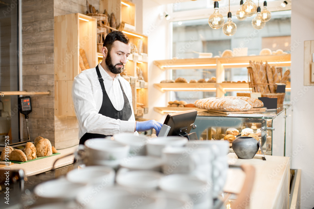 Handsome bread seller working with cash register in the small and cozy store with bakery products Handsome bread seller working with cash register in the small and cozy store with bakery products