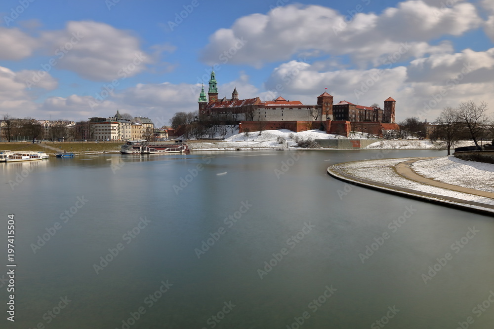 Piękny zimowy pejzaż zakola rzeki Wisły w Krakowie, z zamkiem na wawelu, na rzece statki, błękitne niebo z malowniczymi białymi chmurami, długie naświetlanie